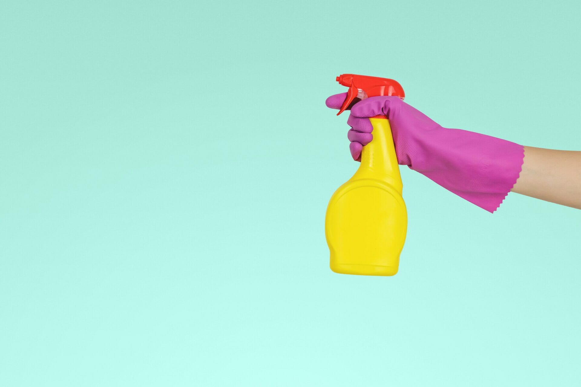 An arm with a pink rubber glove holding a yellow spray bottle against a green background