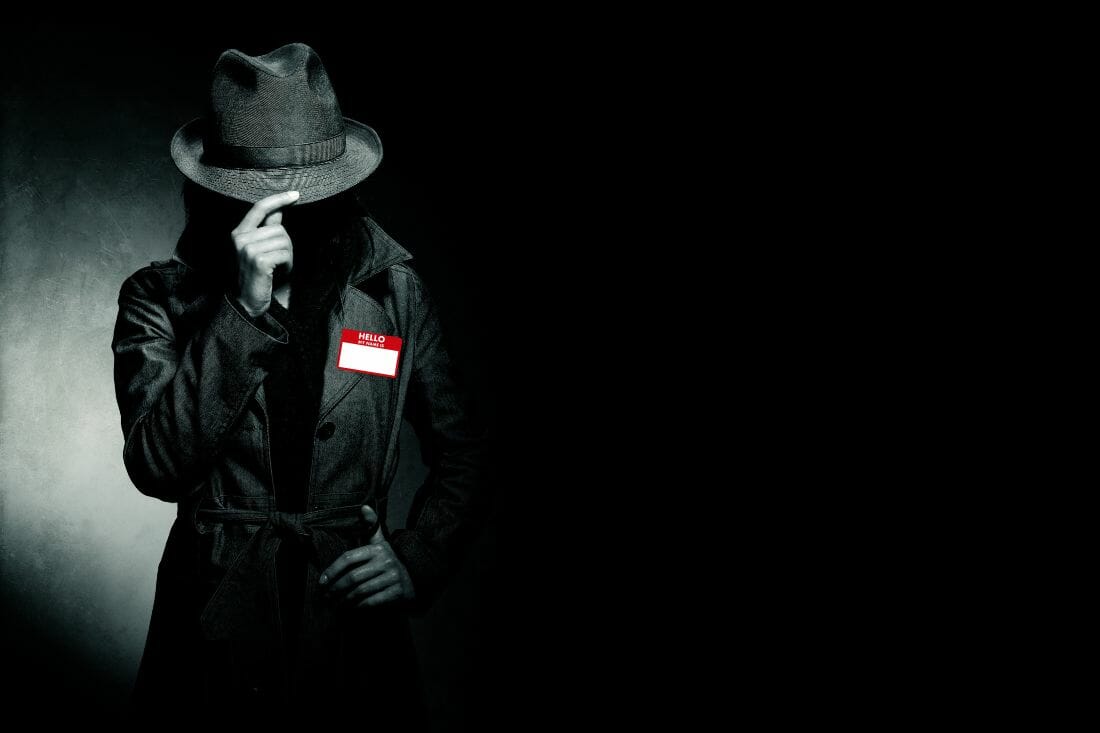 a person in a hat and coat standing against a dark background, the look like a spy. they have a red namebadge memory