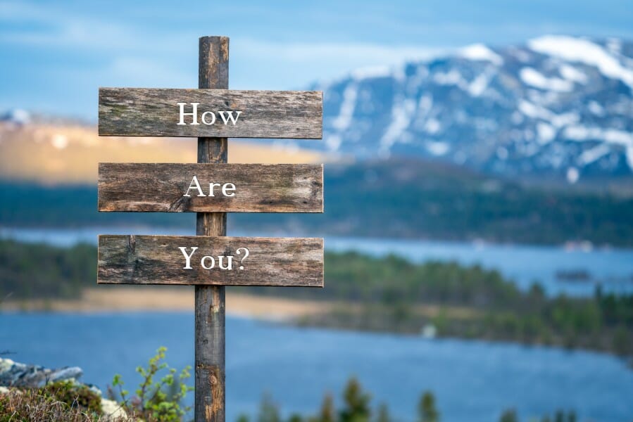 a wooden sign with the words How Are you? in front of a mountain