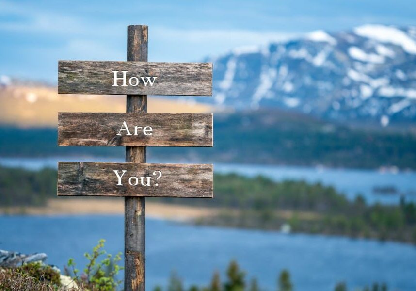 a wooden sign with the words How Are you? in front of a mountain