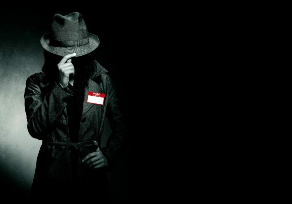 a person in a hat and coat standing against a dark background, the look like a spy. they have a red namebadge memory