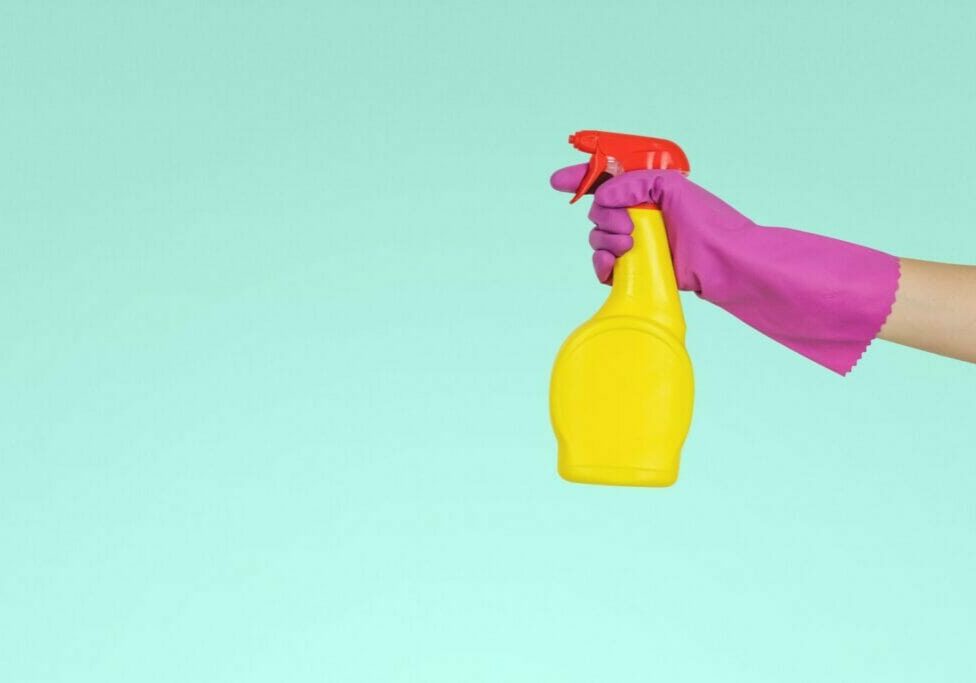 An arm with a pink rubber glove holding a yellow spray bottle against a green background
